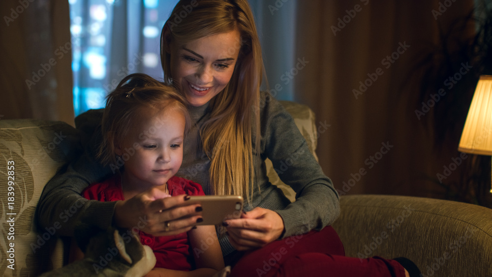 母亲和她可爱的女儿坐在客厅的沙发上。他们正在使用智能手机和