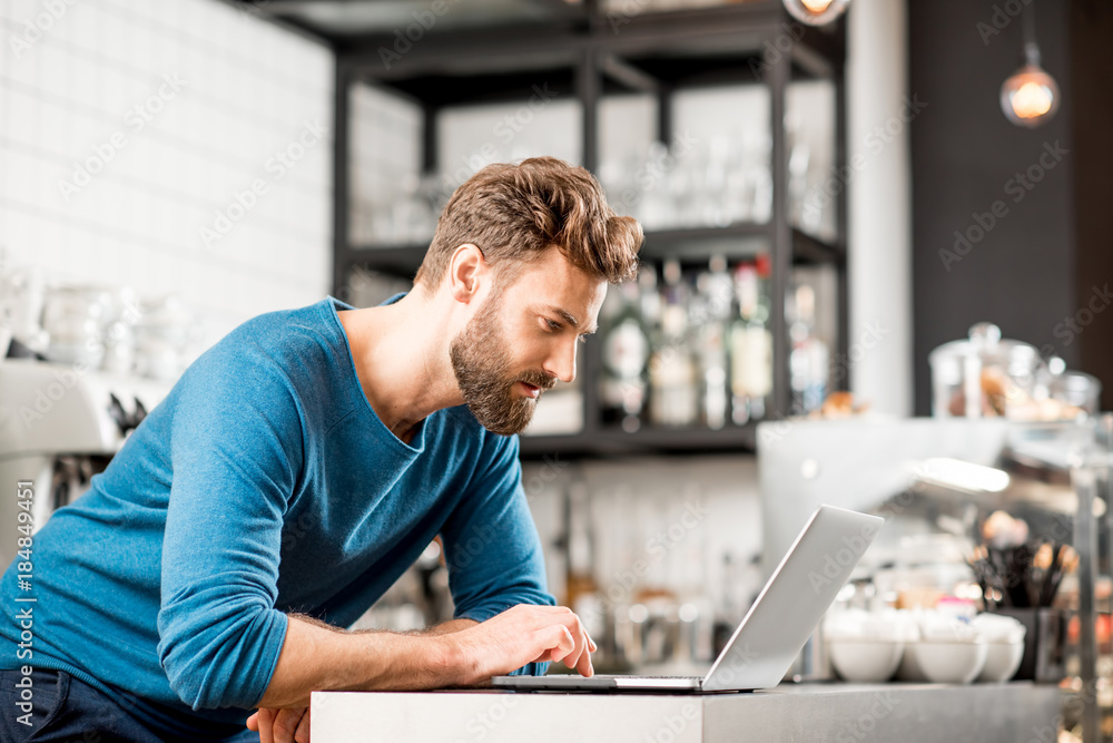 穿着蓝色毛衣的英俊男子在现代咖啡馆内部的酒吧里用笔记本电脑工作