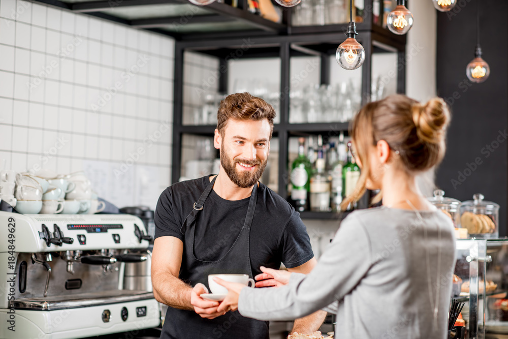 Handsome barista giving a coffee to the female client at the bar of the modern cafe