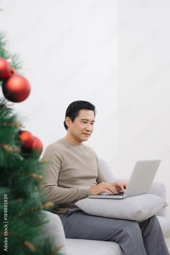 一名男子坐在圣诞树旁的沙发上，拿着笔记本电脑