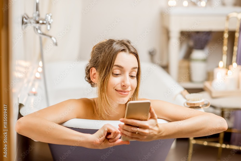 年轻漂亮的女人在室内复古浴缸里用手机放松