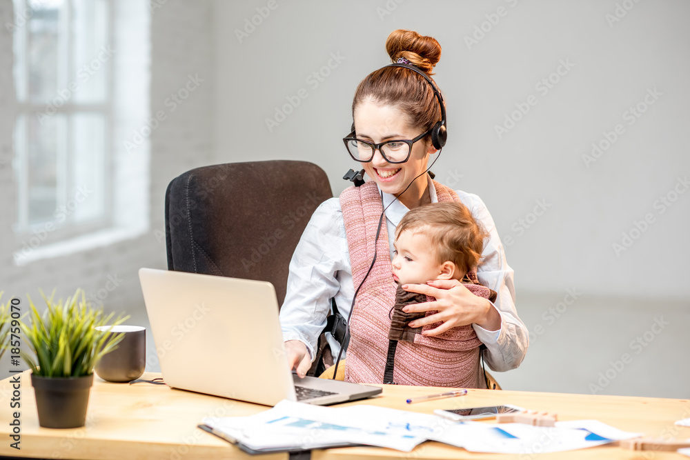 Multitasking businesswoman working with headset and laptop sitting with her baby son at the white of