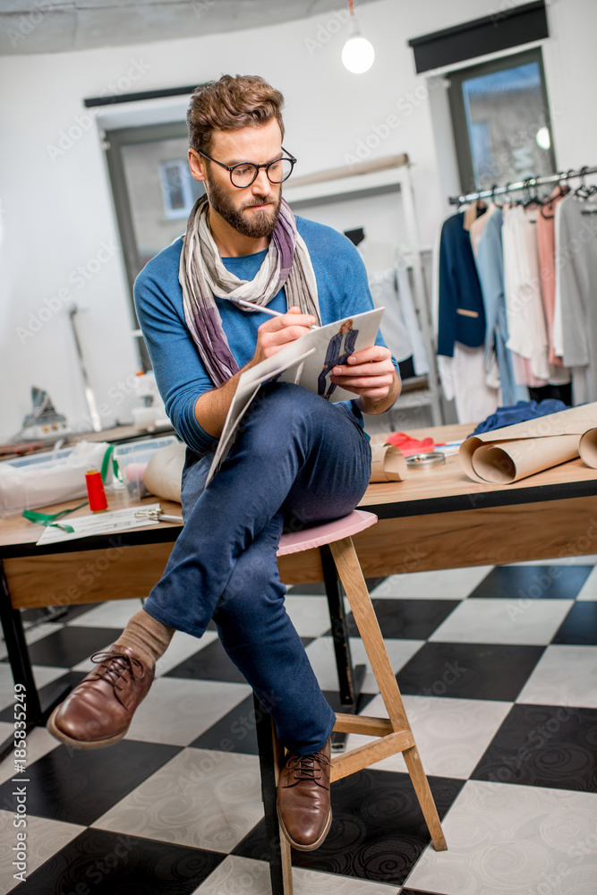 Portrait of a handsome fashion designer sitting with clothing sketches at the studio full of tailori