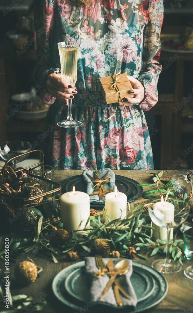 Woman in festive dress holding glass of champaigne and gift box in her hands at holiday decorated ta