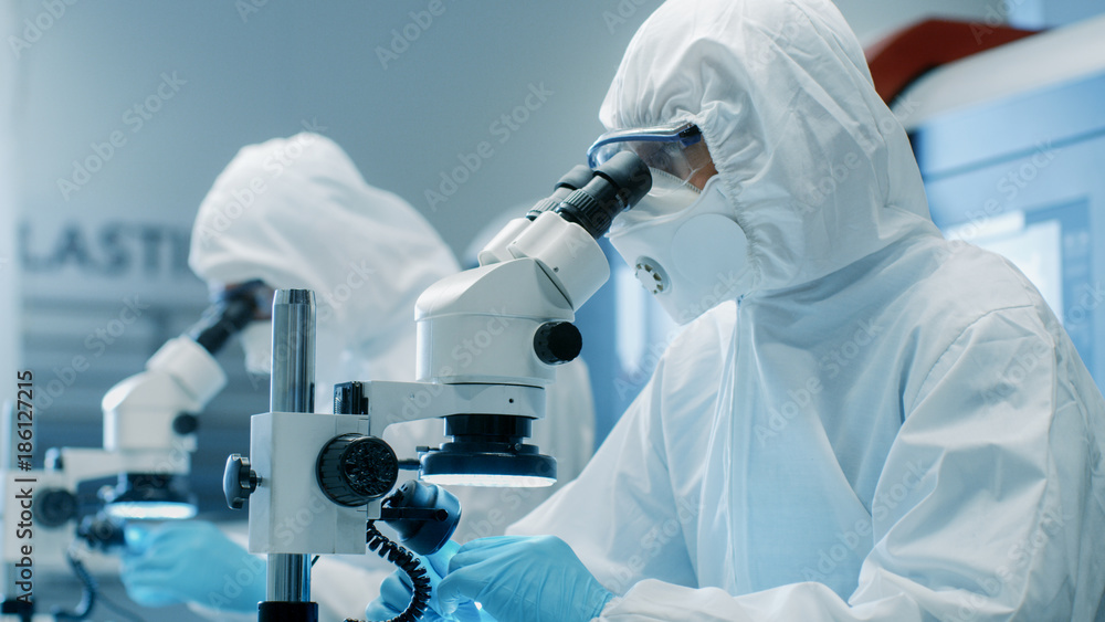 两位科学家在无菌洁净室套装中控制制造机械工作并使用显微镜