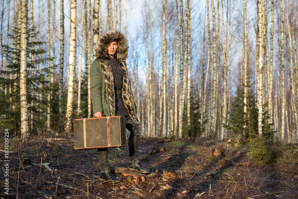 一个穿着绿色冬季派克大衣的年轻人带着行李箱在森林里。