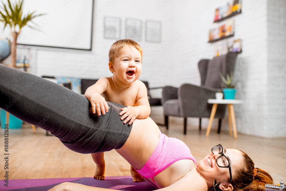 年轻的母亲在垫子上伸展身体，她的小儿子在家里的地板上玩耍