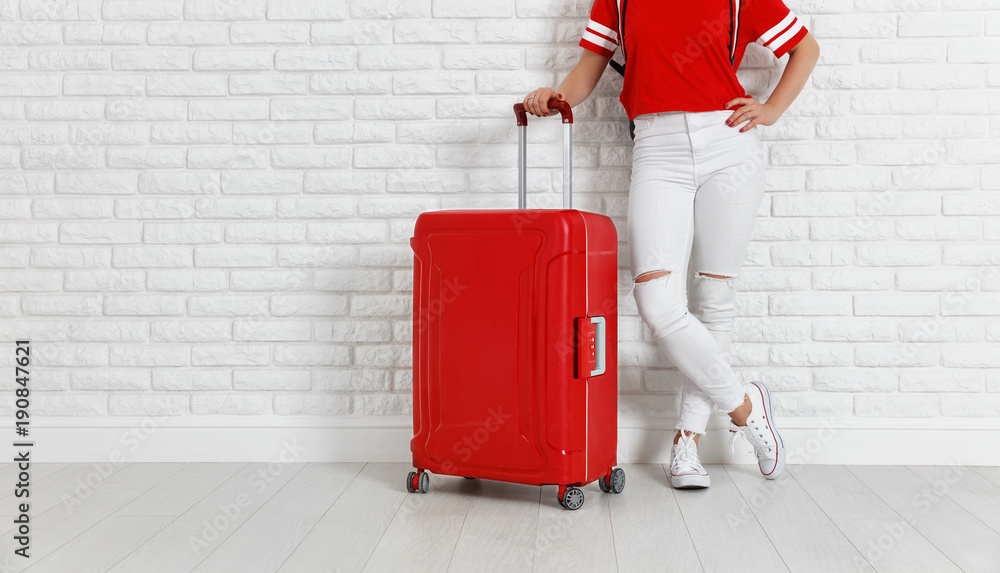 概念旅游。白色空砖墙附近提着红色行李箱的女孩的腿
