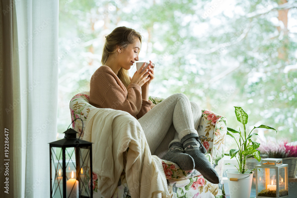 Young woman sitting home in a chair by the window with cup of hot coffee wearing knitted warm sweate