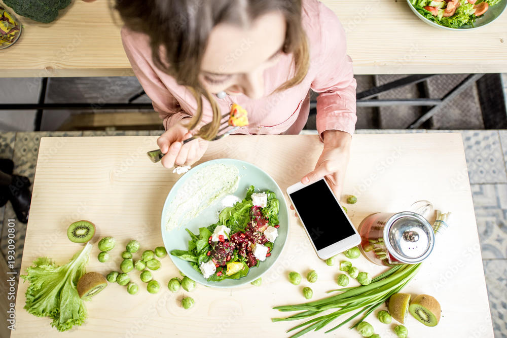 Woman eating salad and using smartphone at the table dwcorated with green food ingredients. Top view