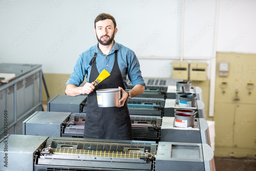 一位英俊的印刷师的肖像，他拿着黄色的颜料站在胶印机的印刷工面前