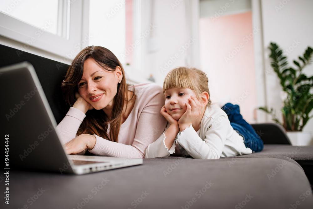 小女孩和她的妈妈躺在沙发上用笔记本电脑看动画片。
