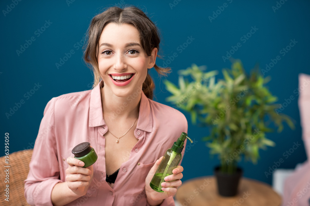 家中蓝色墙壁背景上一位年轻女子拿着装有乳液的绿色瓶子的肖像
