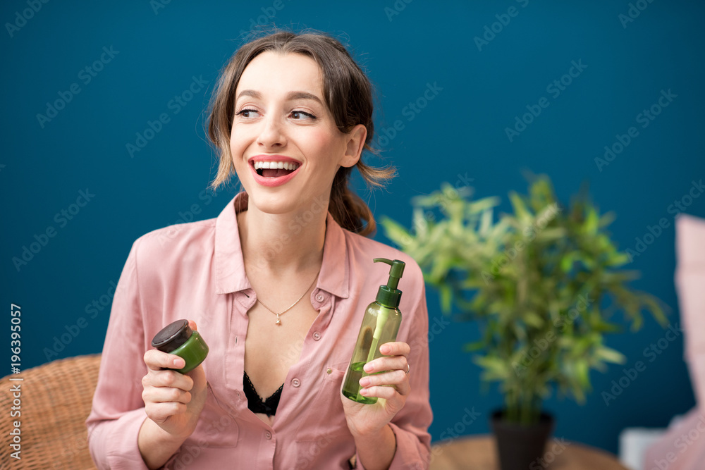 Portrait of a young woman holding green bottles with lotion on the blue wall background at home