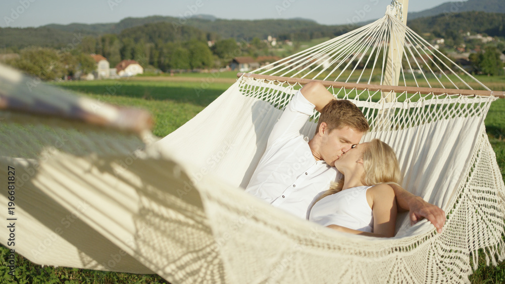 Young Caucasian woman and handsome man cuddle and kiss in romantic hammock.