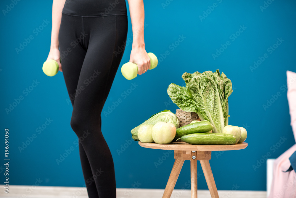 蓝色墙壁背景上有哑铃和健康绿色食物的运动女性身体