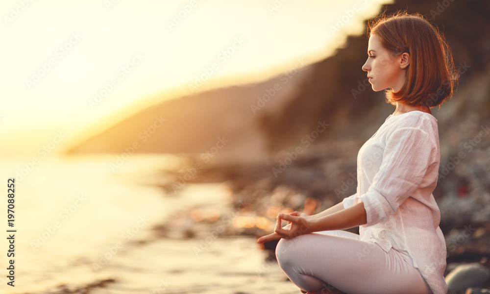 女人在日落海滩上练习瑜伽并以莲花姿势冥想。