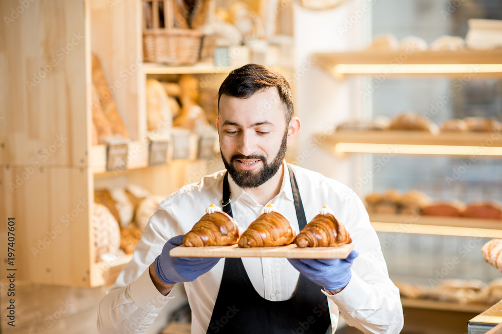 一个穿着制服的英俊卖家拿着美味的羊角面包站在面包店的画像
