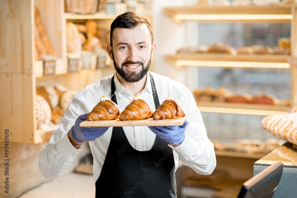 一个穿着制服的英俊卖家拿着美味的羊角面包站在面包店的画像