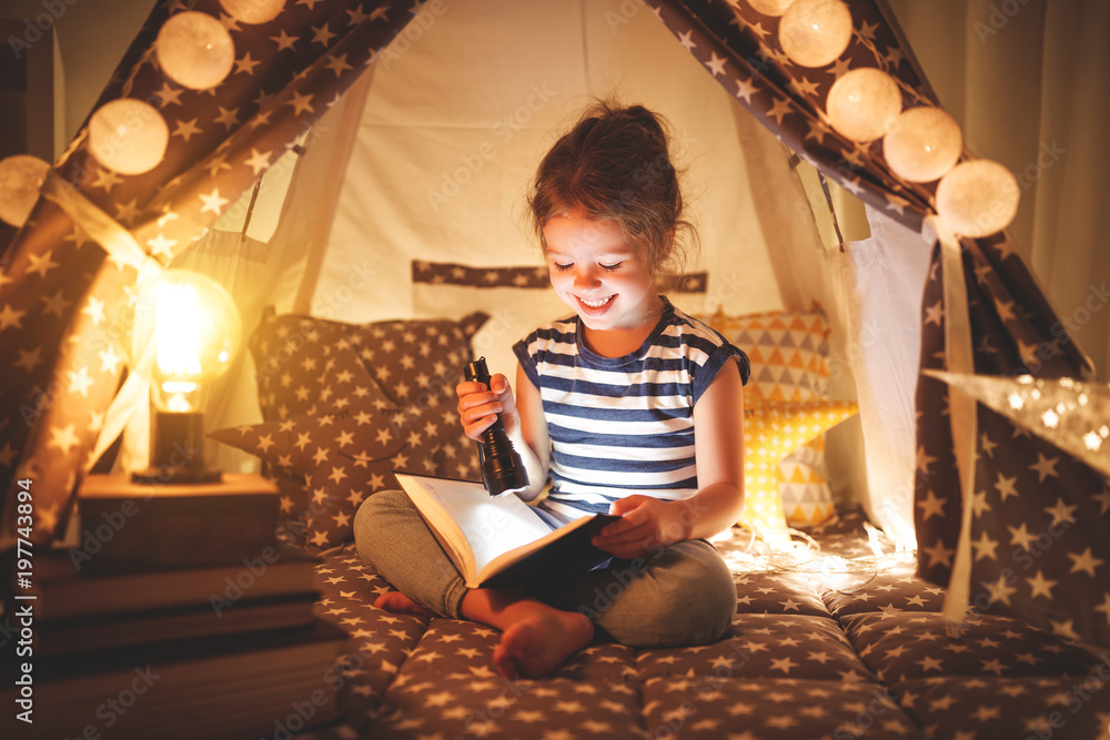 快乐的小女孩在家里的帐篷里，在黑暗中笑着看书