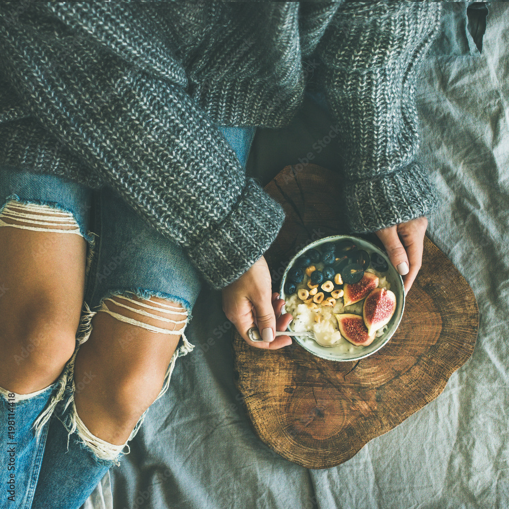 床上的健康冬季早餐。穿着毛衣和牛仔裤的女人在吃无花果椰子粥，
