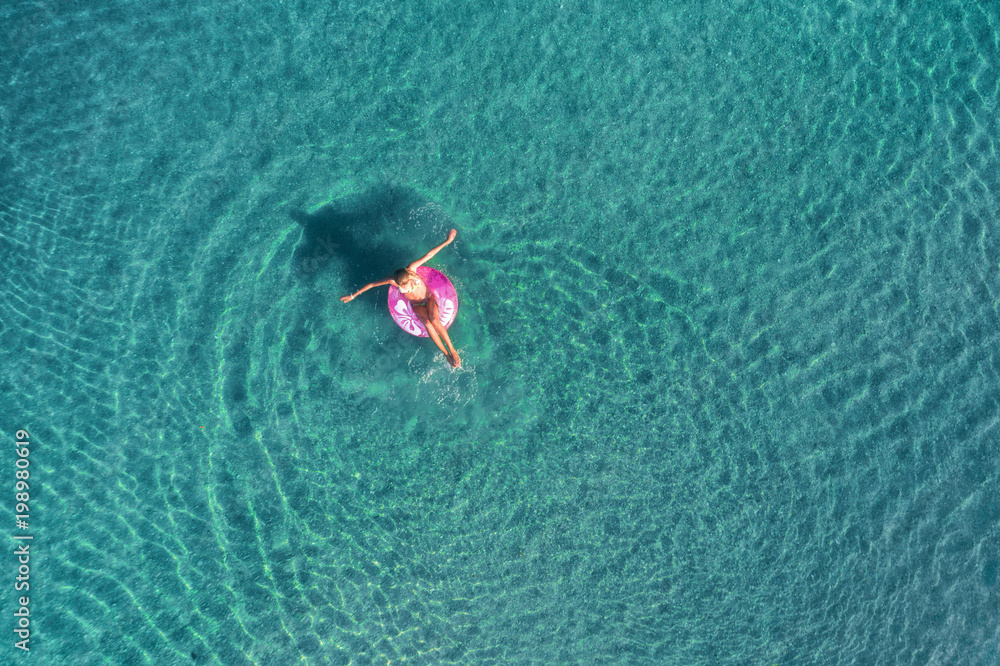 日出时，一名年轻女子在透明的绿松石海中的粉色游泳圈上游泳的鸟瞰图