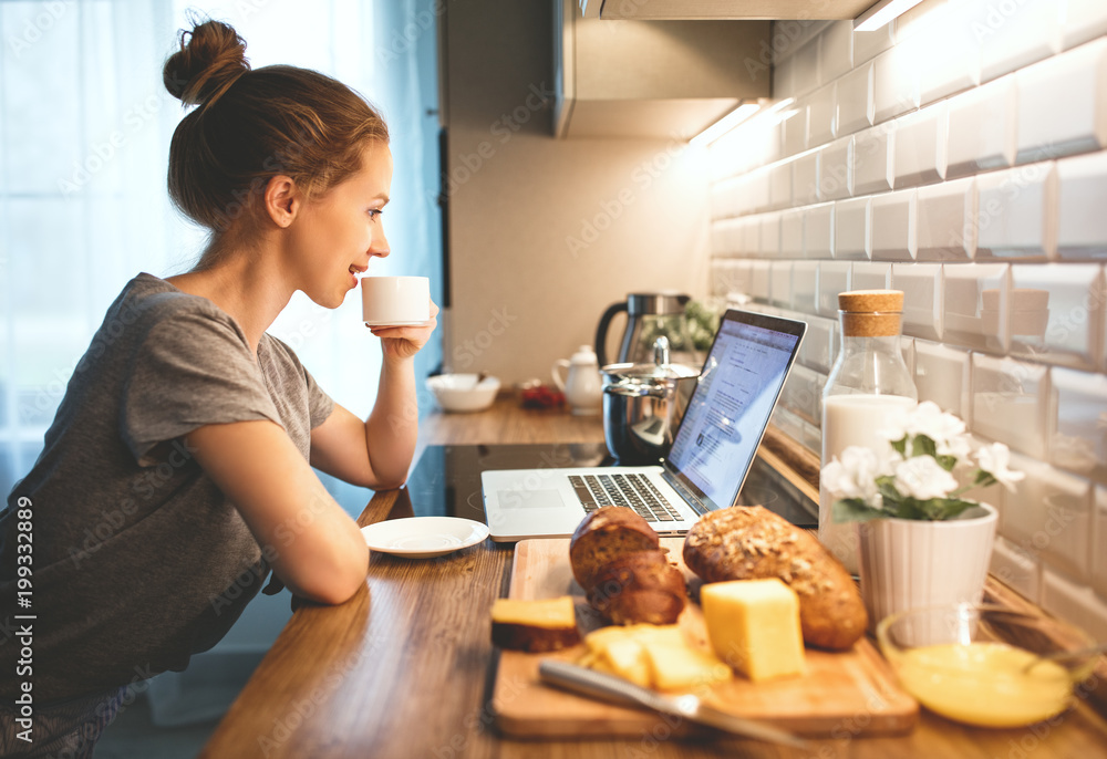 woman in pajamas is having breakfast at computer in  kitchen.