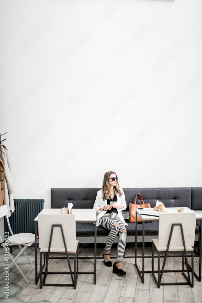 穿着白色夹克的时尚商务女性，在现代咖啡馆内部的咖啡休息时间坐着。