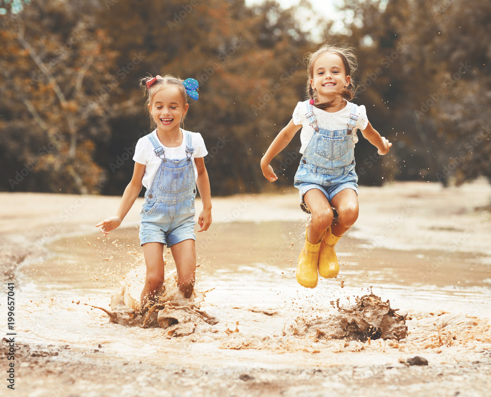 快乐有趣的姐妹双胞胎小女孩穿着橡胶靴在水坑上跳跃。