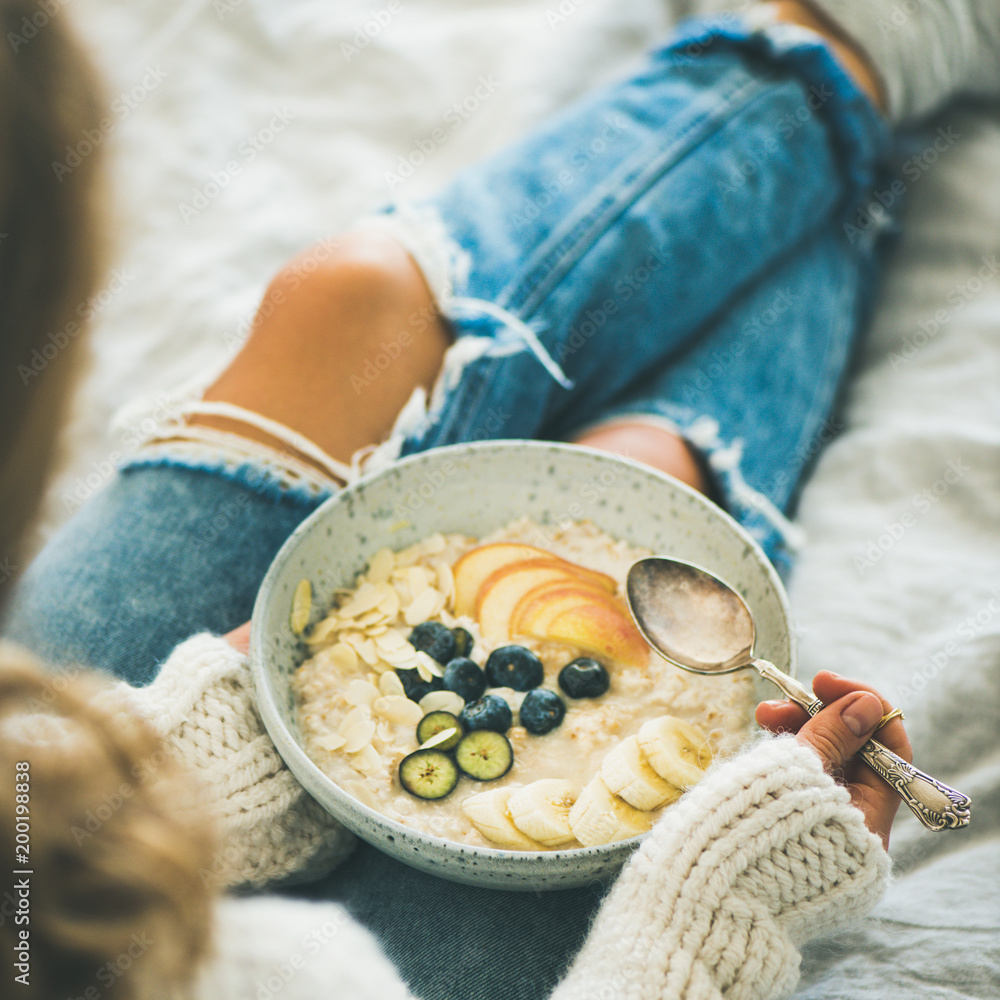 健康的冬季早餐在床上。穿着羊毛衫和破旧牛仔裤的女人在吃纯素食杏仁奶o