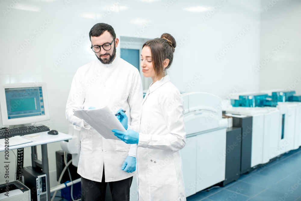 两名医护人员拿着纸质文档站在实验室里，上面有分析仪