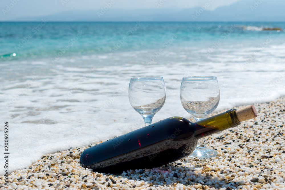 夏日阳光明媚的海滩上，一瓶带着酒杯的红酒。背景是大海。