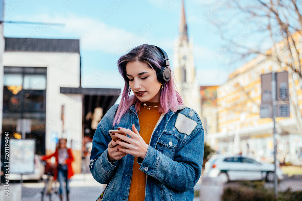 漂亮的女孩，紫色头发，戴着耳机，在城市街道上。看着手机。