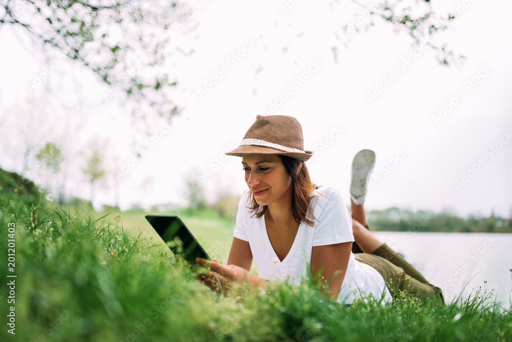 在户外使用平板电脑的年轻女性躺在草地上，面带微笑。