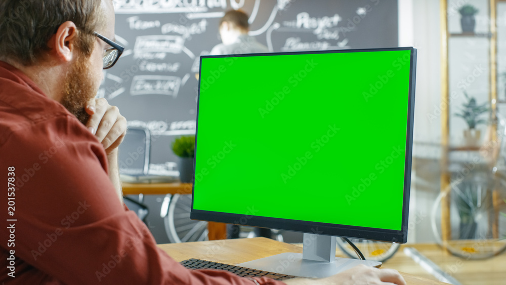 男人在带绿色实体屏幕的个人电脑上工作。在后台创意办公室里