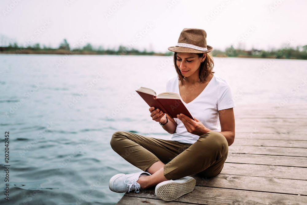美丽的女人坐在木码头上看书。