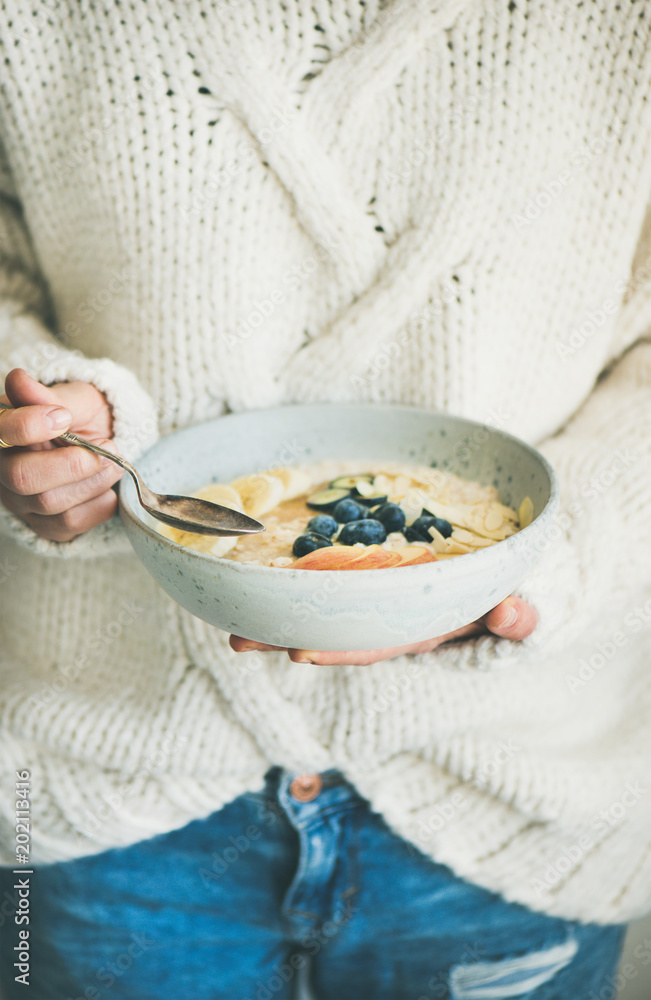 Healthy winter breakfast in bed. Woman in woolen sweater and jeans eating vegan almond milk oatmeal 