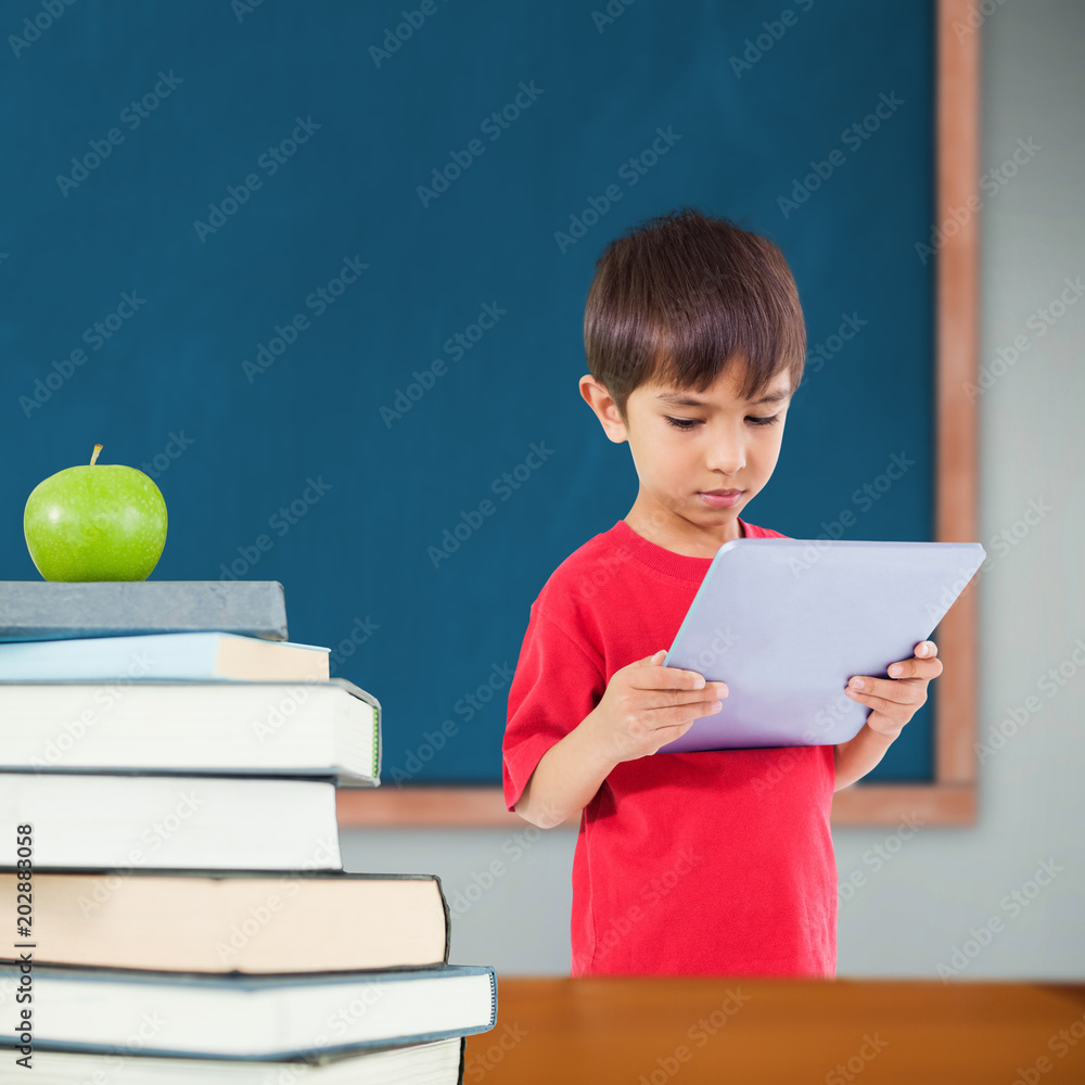 Cute boy using tablet against green apple on pile of books in classroom