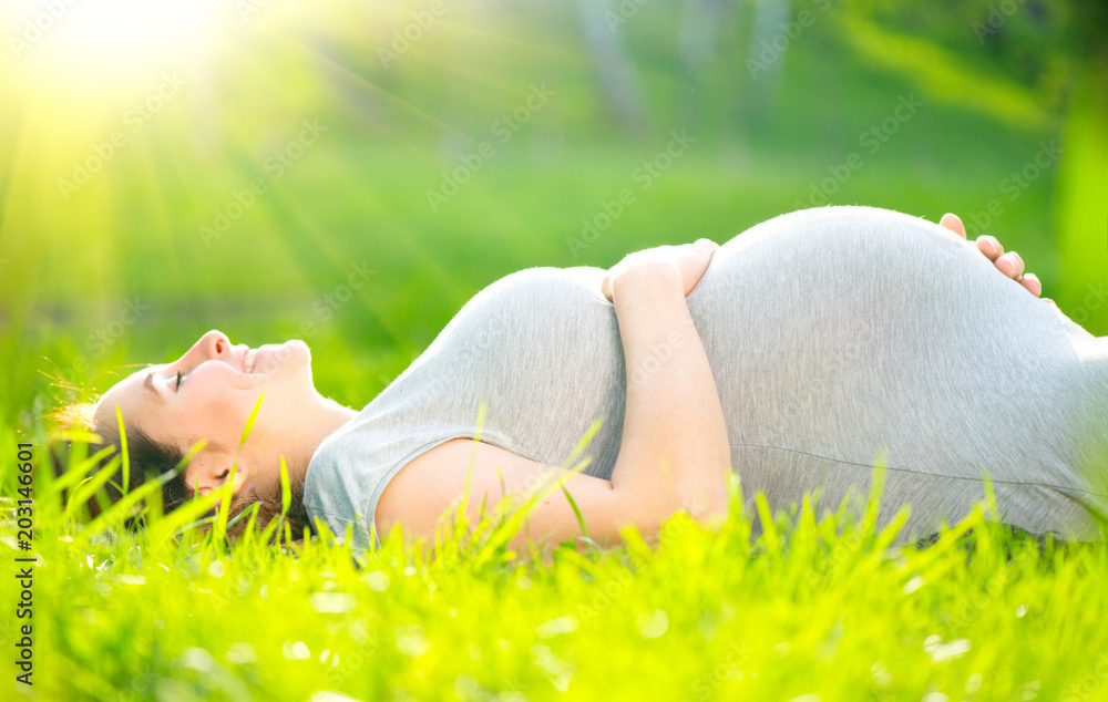 怀孕的中年妇女躺在绿草上抚摸腹部，享受大自然，健康怀孕