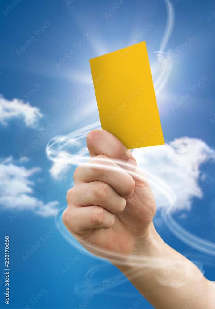 Hand holding up yellow card against bright blue sky with clouds
