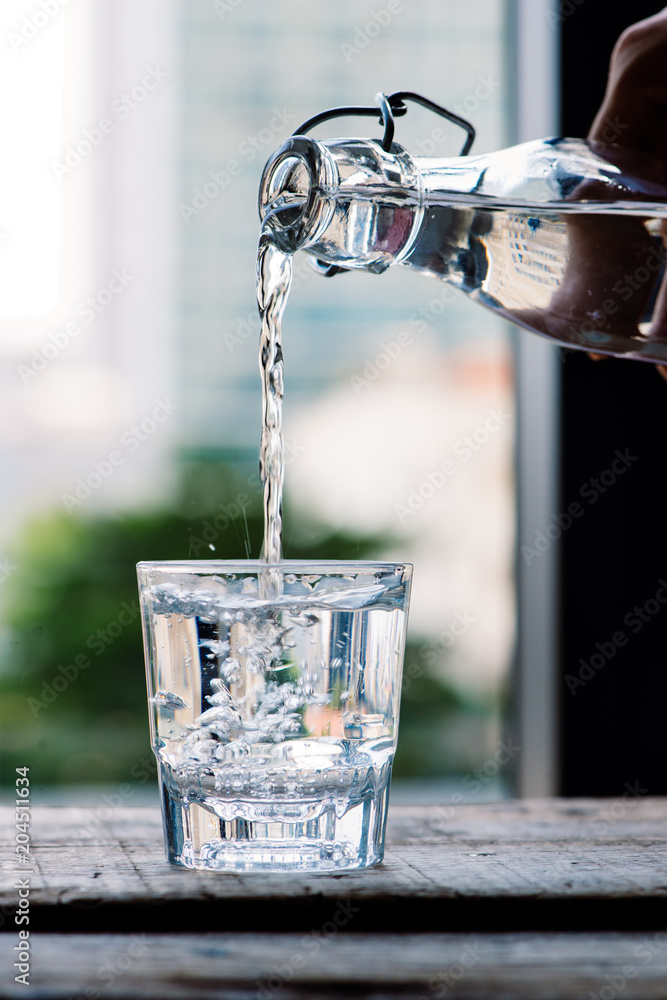 干净的饮用水从一个罐子里倒进一个木制桌子上的圆形玻璃杯和一个浅绿色