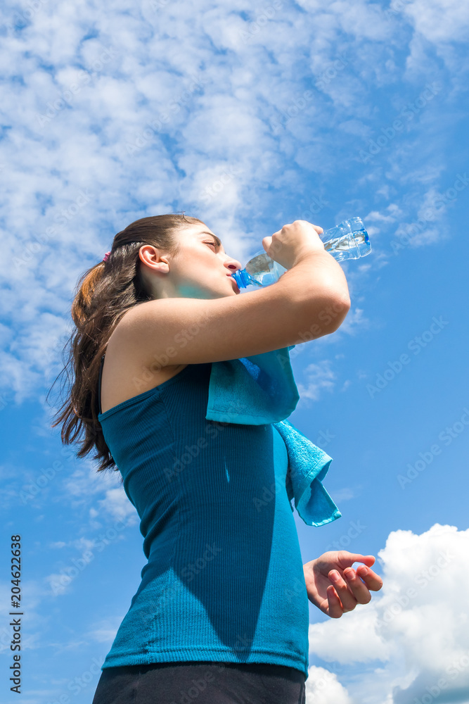 美丽的女跑步者正在休息，在晴朗的蓝天背景下喝水