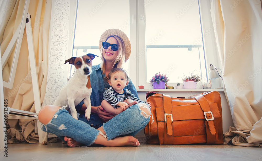 带着小婴儿和一只狗的年轻女孩坐在行李箱旁边，然后在家里的窗户附近旅行