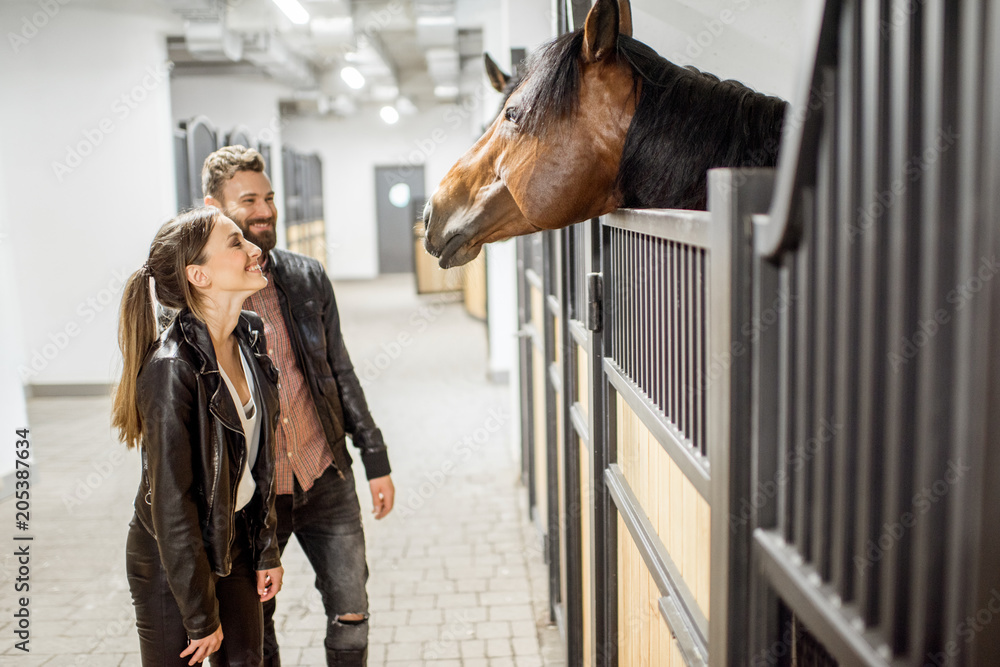 穿着皮夹克的年轻情侣骑手与马站在美丽的马厩里