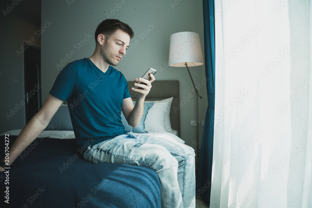 英俊的高加索男子在晨光中用智能手机联系卧室里的人