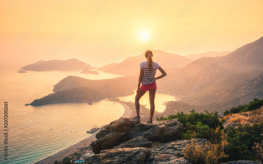年轻的女人站在岩石的顶部，看着五颜六色的日落中的海岸和山脉