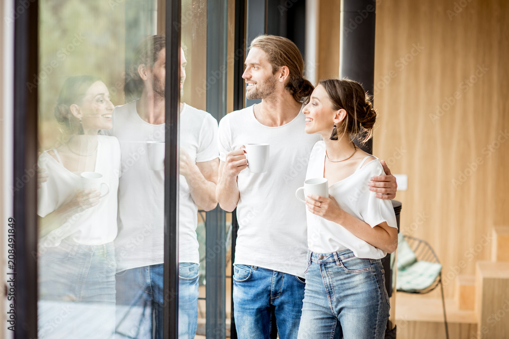 一对年轻幸福的夫妇穿着白衬衫，端着杯子站在家里的窗户边