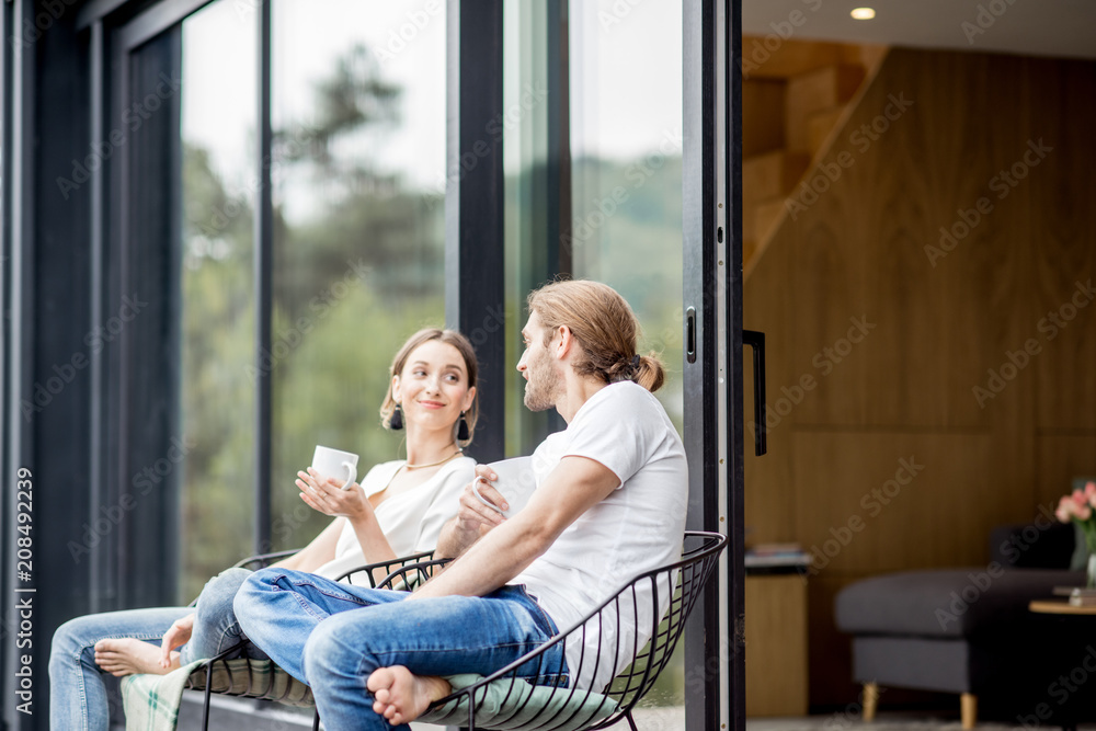 年轻夫妇端着杯子坐在现代房子的露台上，在户外交谈