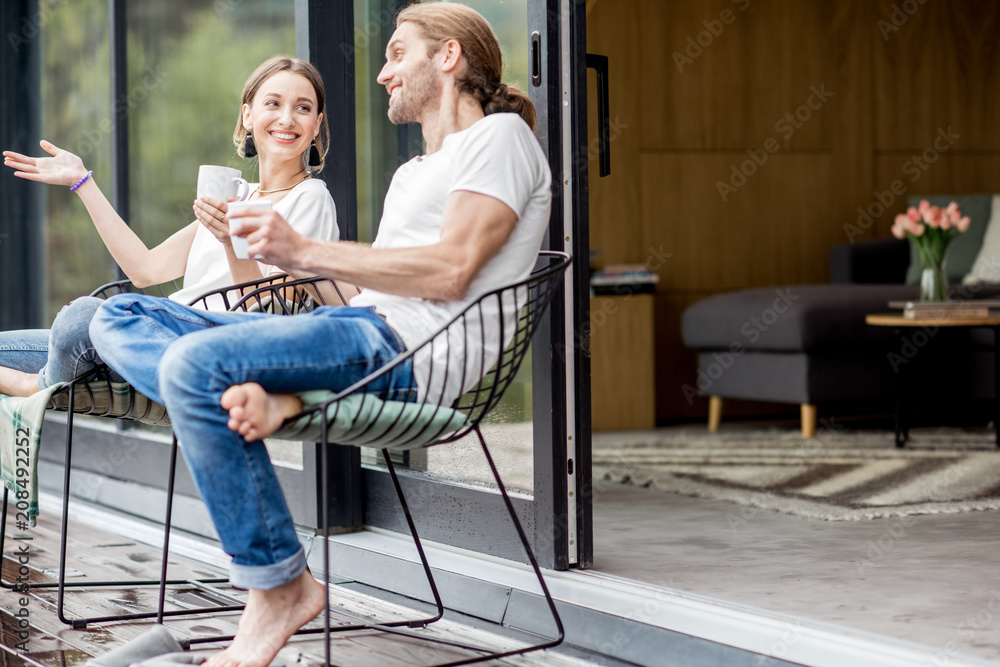 Young couple sitting with cups on the terrace of the modern house talking together outdoors