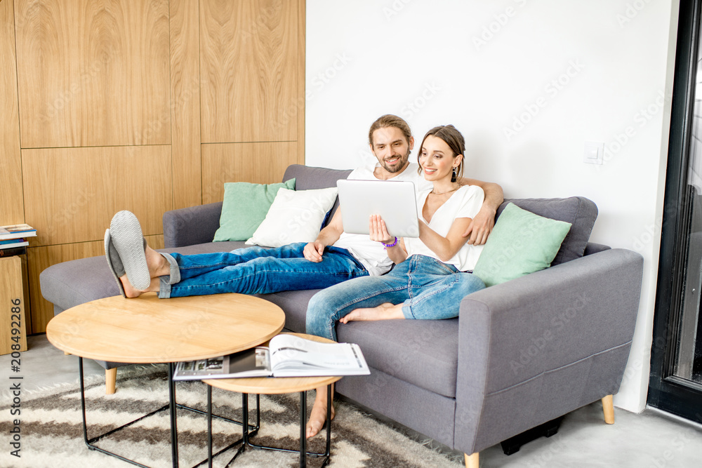 一对年轻夫妇穿着白衬衫和牛仔裤，使用数字平板电脑坐在沙发上